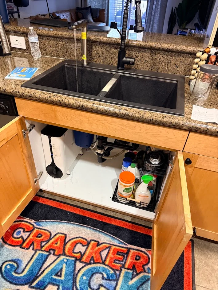 A double-basin kitchen sink with a tall black faucet above an open wooden cabinet showing plumbing, containers and cleaning supplies.