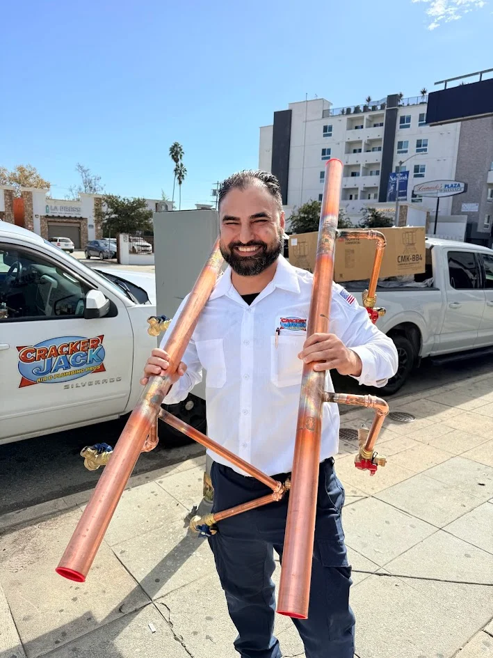 A person in a white shirt holding two large copper pipe assemblies outdoors, with parked vehicles and buildings behind them.