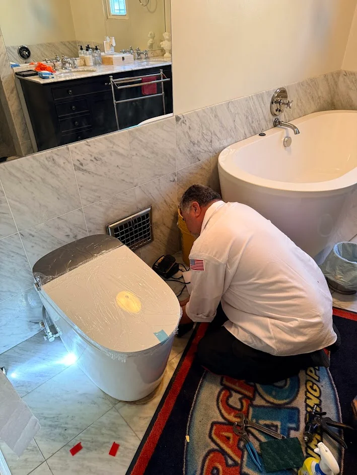 A person kneeling beside a toilet with tools on a floor mat in a bathroom.