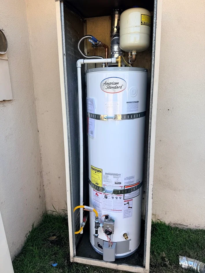 Cylindrical water heater secured with metal straps inside an open utility cabinet, with connected piping and a small tank above.