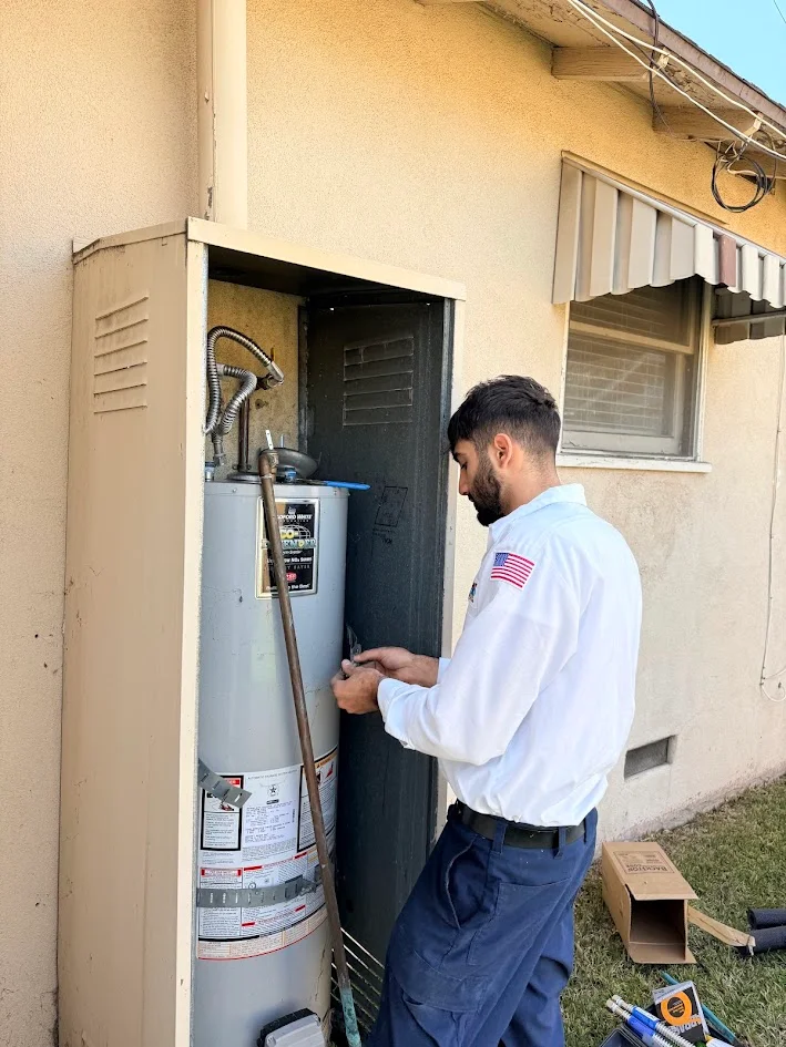 A person holds a small tool beside a cylindrical water heater inside an open exterior cabinet. Tools and an open cardboard box sit nearby.