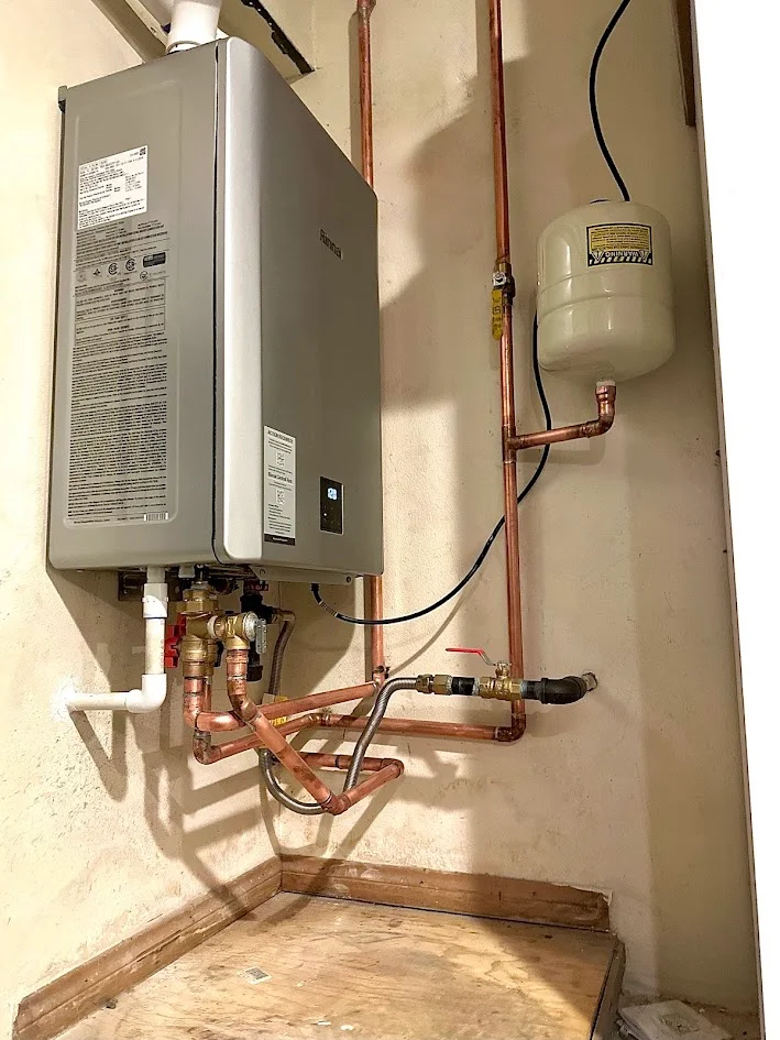 Wall-mounted water heater connected to copper pipes, valves and a small rounded tank above a wooden platform.