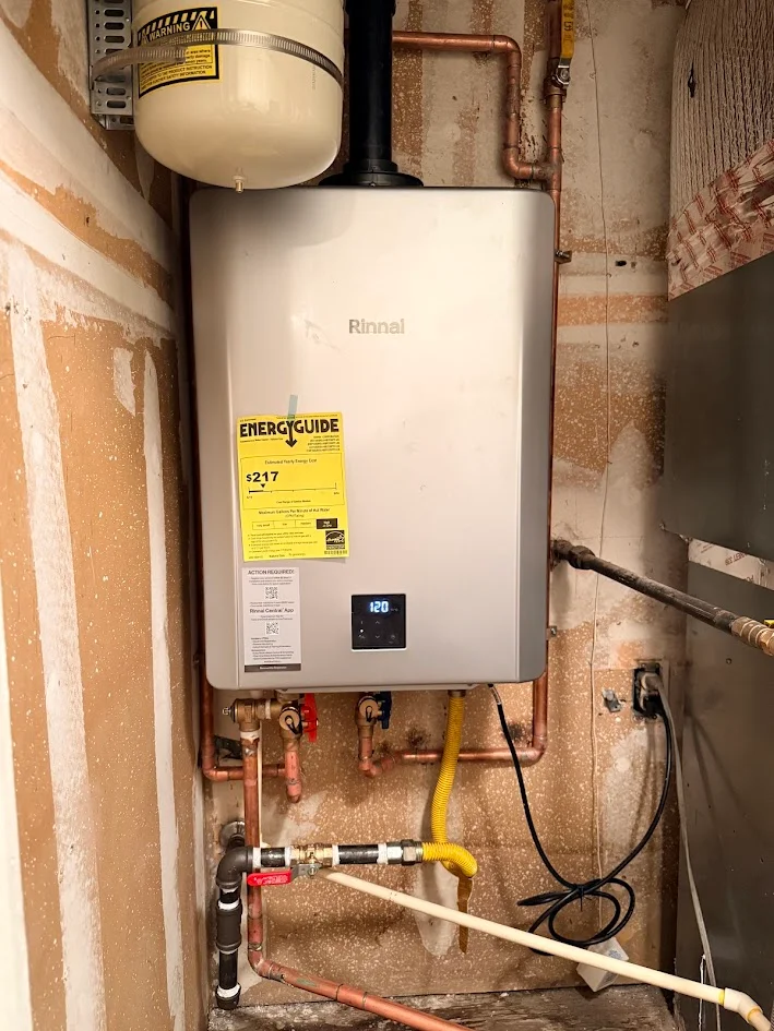 Wall-mounted water heater with an expansion tank, connected pipes, valves and electrical cord in an unfinished utility space.